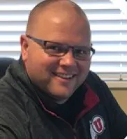 Smiling bald man with glasses wearing a dark jacket with a University of Utah logo inside a room with window blinds.