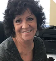 Smiling woman with curly dark hair wearing a gray top sitting indoors near office equipment.