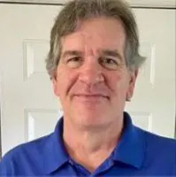 Middle-aged man with gray hair wearing a blue polo shirt smiling in front of a white door background