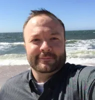 Bearded man wearing a button-up shirt taking a selfie at a sunny beach with waves and blue sky.