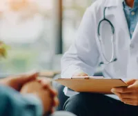 Doctor in white coat with stethoscope holds clipboard during patient consultation in clinic.
