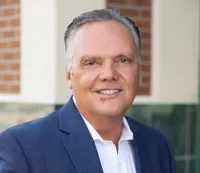 Smiling middle-aged man in a blue suit and white shirt standing outdoors with a blurred brick and tile background.