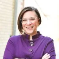 Smiling woman with short hair and glasses wearing a purple buttoned shirt with arms crossed.
