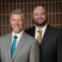 Two men in business suits smiling, one with gray hair and teal tie, the other bald with beard and gold tie.