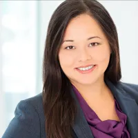 Professional woman with long dark hair smiling, wearing a dark blazer and purple blouse in bright office setting