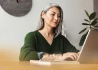 Middle-aged woman with gray hair working on a laptop at a wooden desk with glasses and clock in background