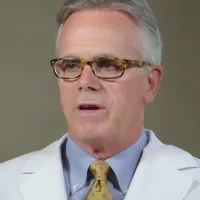 Mature male doctor wearing glasses, white coat, yellow tie, speaking in a professional setting with plant background