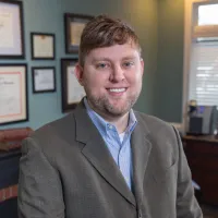 Smiling man in business attire standing in an office with framed certificates and a window with blinds.