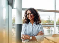 Smiling woman with curly hair and glasses standing in bright modern office with arms crossed.