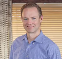 Smiling man in a blue checkered shirt standing indoors with window blinds and patterned curtains behind him.