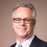Smiling middle-aged man with gray hair, wearing tortoiseshell glasses, a suit, white shirt, and striped tie