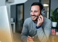 Smiling man wearing headset working on computer in modern office setting with plants and shelves in background