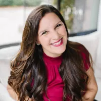 Smiling woman with long brown hair wearing a burgundy top sitting on a white couch indoors.