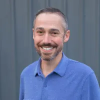 Smiling man with short hair and beard wearing a blue collared shirt against a gray background