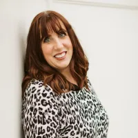 Smiling woman with red hair wearing a black and white leopard print top leaning against a white wall