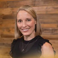 Smiling woman with shoulder-length blonde hair wearing a black top and heart necklace against a wooden background