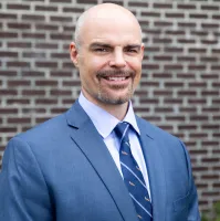 Smiling bald man in blue suit and patterned tie standing in front of a brick wall outdoors.
