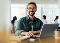 Smiling man with glasses working on laptop at desk with notebook and coffee in modern office space.