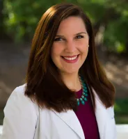 Smiling woman with long brown hair wearing a white coat and turquoise necklace outdoors in natural light