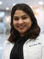 Smiling female dentist in white coat with Aparna Menon, DMD embroidered, in bright dental office.
