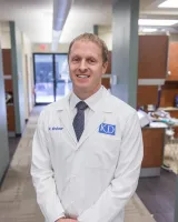 Smiling male doctor in white coat and tie standing in a modern medical office hallway.