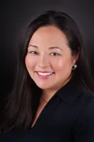 Smiling woman with long black hair wearing a dark blouse and silver hoop earrings against a black background