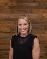Smiling blonde woman in a black sleeveless top with heart necklace, standing against a wooden plank wall background.
