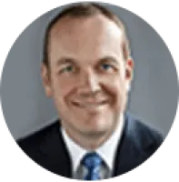 Professional headshot of a smiling man in a suit and blue tie with a neutral gray background