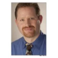 Portrait of a smiling man with red hair, beard, blue shirt, and patterned tie against a neutral background.