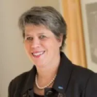 Smiling woman with short gray hair, wearing pearls and a dark blazer, in a professional setting.