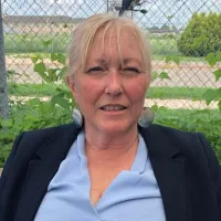 Blonde woman in blue shirt and black blazer sitting outdoors with greenery and a fence in the background