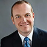 Professional portrait of a smiling man in a suit with a blue tie against a gray background.
