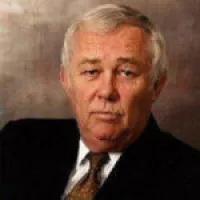Portrait of a senior man with gray hair in a dark suit and patterned tie against a brown background.