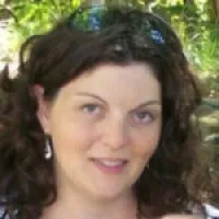 Smiling woman with curly brown hair and dangling earrings outdoors with green foliage background