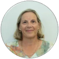 Smiling middle-aged woman with short blonde hair wearing a multicolored top and gold jewelry against white background.