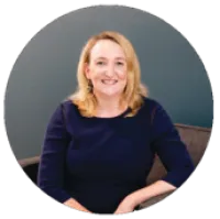 Smiling blonde woman in navy blue top sitting on a chair against a gray background