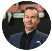 Man in blue blazer and black shirt standing in front of a blurred company logo background.