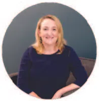 Blonde woman in dark blue top sitting on a chair with a neutral gray background, smiling at the camera.