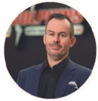 Man in dark blazer and black shirt standing in front of a blurred background with red and white text.