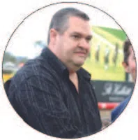 Middle-aged man in a dark checkered shirt standing outdoors with a blurred green background.