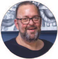 Middle-aged man with glasses and beard smiling against a blue and patterned background.