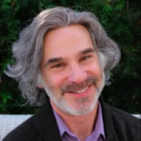 Smiling man with long gray hair, wearing a purple shirt and dark blazer, outdoors with greenery in the background.