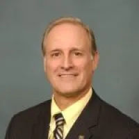 Professional headshot of a businessman wearing a suit and tie, smiling against a muted background.
