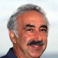 A smiling man with gray hair and mustache against a blurred outdoor background.