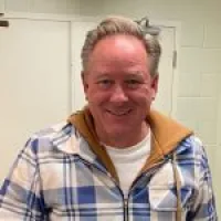 Smiling man in a plaid shirt and tan jacket, standing in front of a light-colored wall.