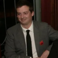 A man in a gray suit and black tie smiling in an elegant setting with a glass in front of him.