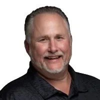 Smiling middle-aged man with short gray hair and beard wearing a black patterned shirt against white background