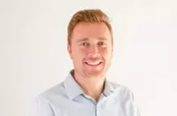 Smiling young man with light skin and blonde hair wearing a light blue collared shirt against a plain background