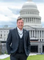 Smiling man in a dark suit with beaded necklaces stands in front of the U.S. Capitol building on a cloudy day.