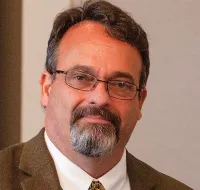 Middle-aged man with glasses and a beard wearing a brown suit jacket and patterned tie against a neutral background